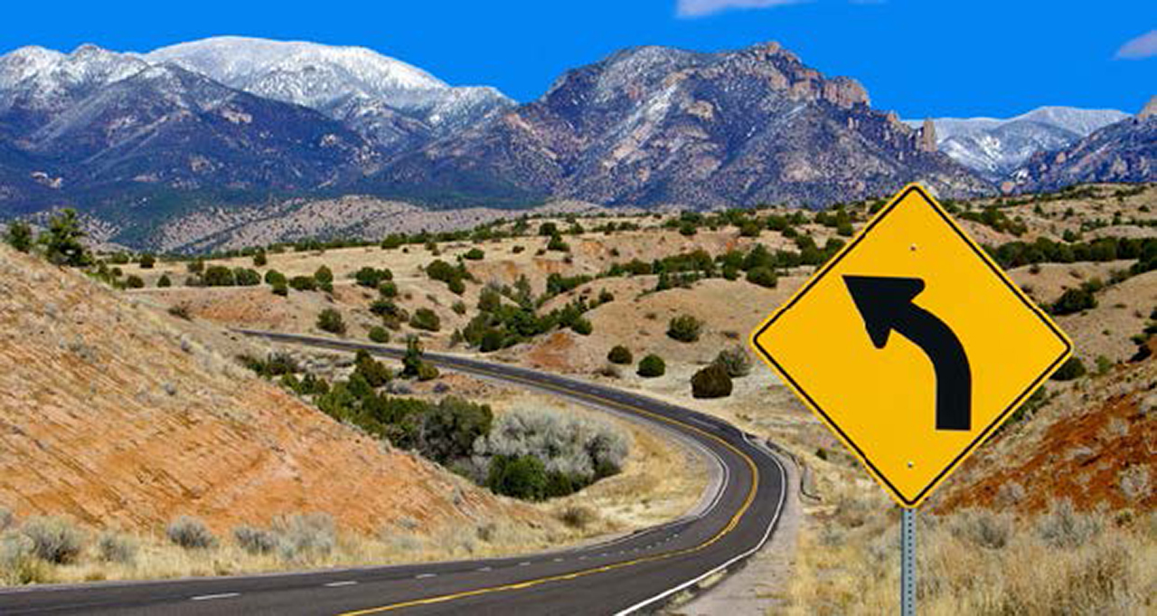 photo of a curved highway in the desert with snow-capped mountains in the background. Alongside the roadway is a yellow warning sign containing a curved arrow