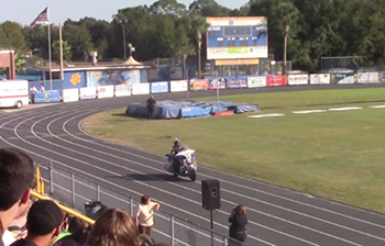 Screen capture from the safety video shows a police officer riding a motorcyle on the track around a high school field while students watch from the stands